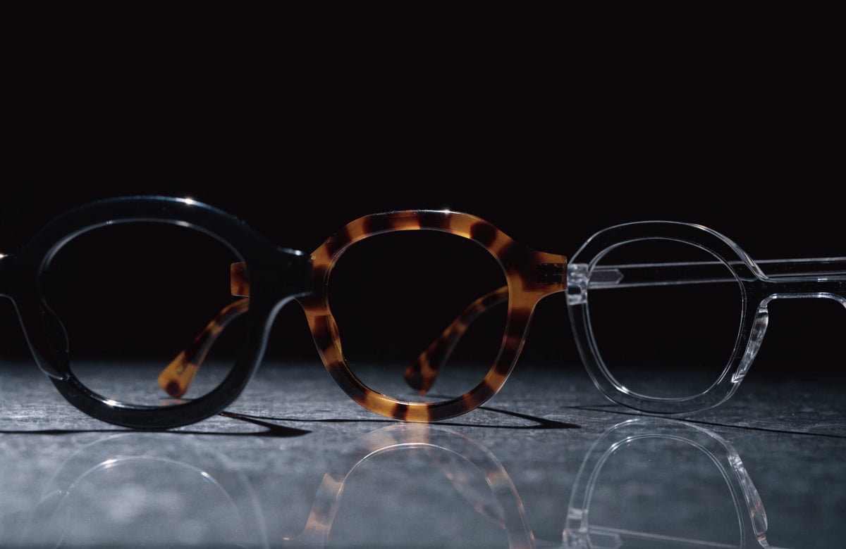 Three independent luxury acetate eyewear frames in black, tortoiseshell and clear on a dark stone surface under hard light
