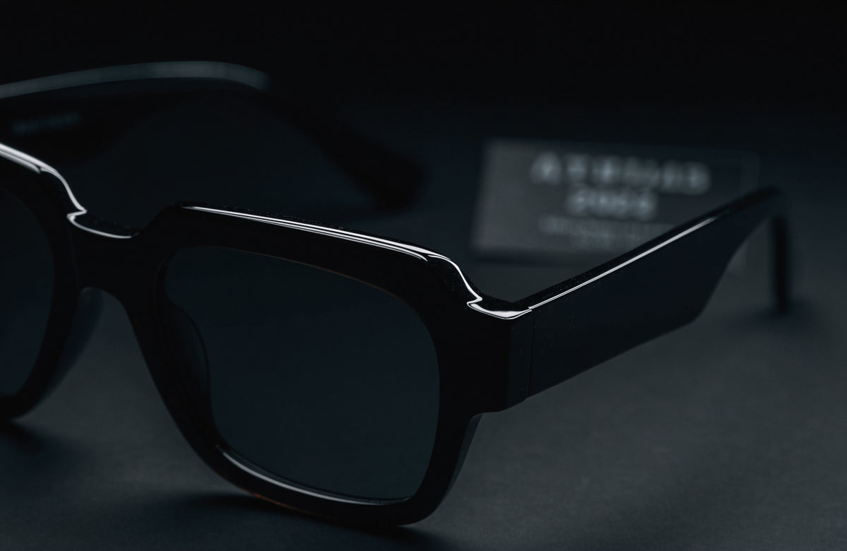Black acetate sunglasses on a dark surface with a numbered edition certificate card blurred in the background