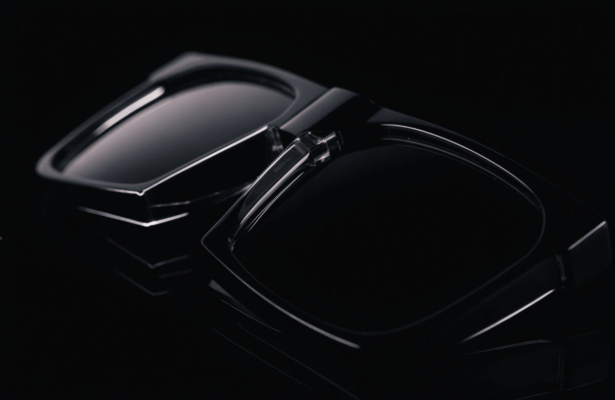 Black folding sunglasses partially collapsed at the bridge hinge on a black surface under hard studio light