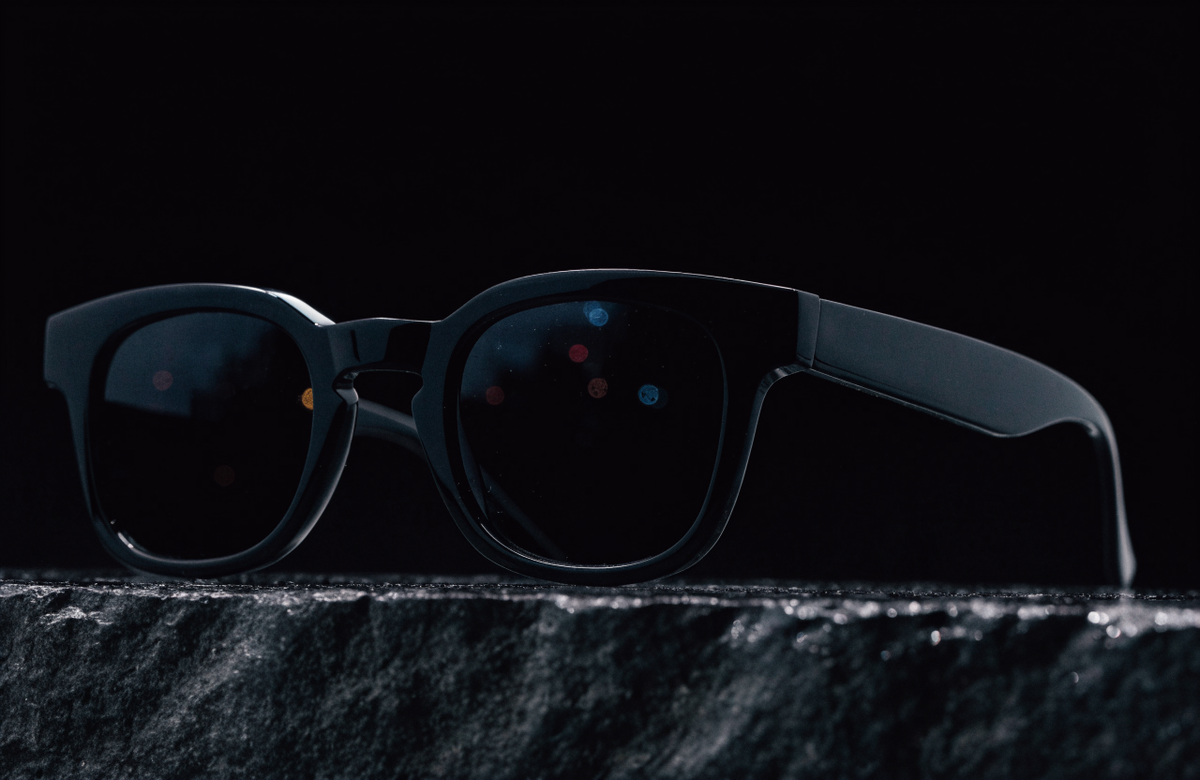 Black acetate sunglasses on a dark stone ledge at night with city lights reflected in the lenses