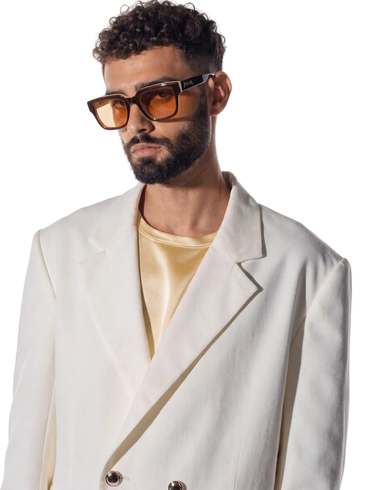 A man with a beard and curly hair wearing oversized square sunglasses with amber-tinted lenses and dark brown frames, styled with a cream blazer over a pale yellow satin shirt, against a plain white studio background.