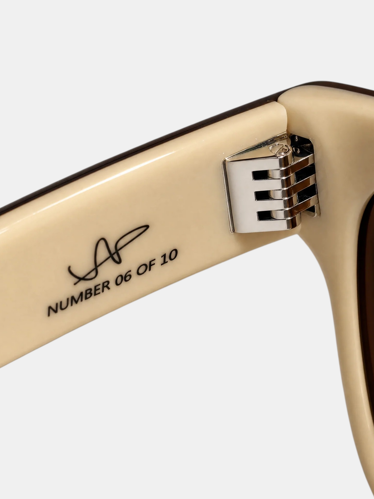 Close-up of a cream-colored sunglasses arm with black printed text reading 'NUMBER 06 OF 10' and a stylized signature logo, plus a silver metal hinge detail, on a light gray background. PULERA